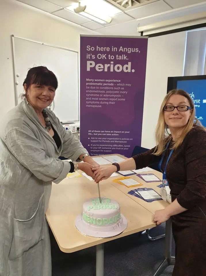 Launch of the Angus Council Periods and Menopause Policy. Image shows Jools Bell cutting a cake, across the table from a colleague. A poster behind her describes problematic period symptoms and encourages women to talk about it.